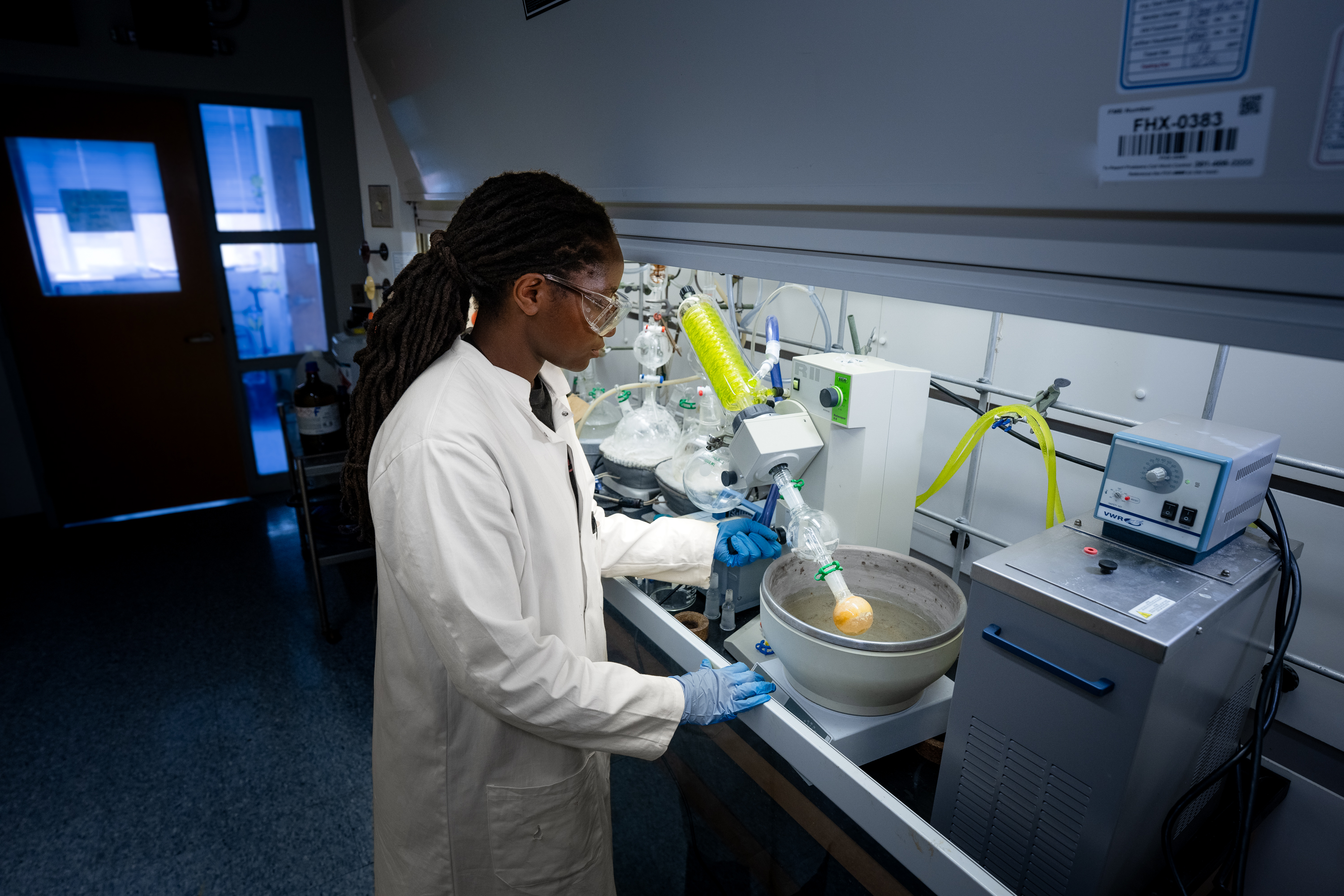 A researcher in a lab coat, safety glasses, and gloves operates round device in a fume hood, with a round-bottom flask submerged in a heated water bath.