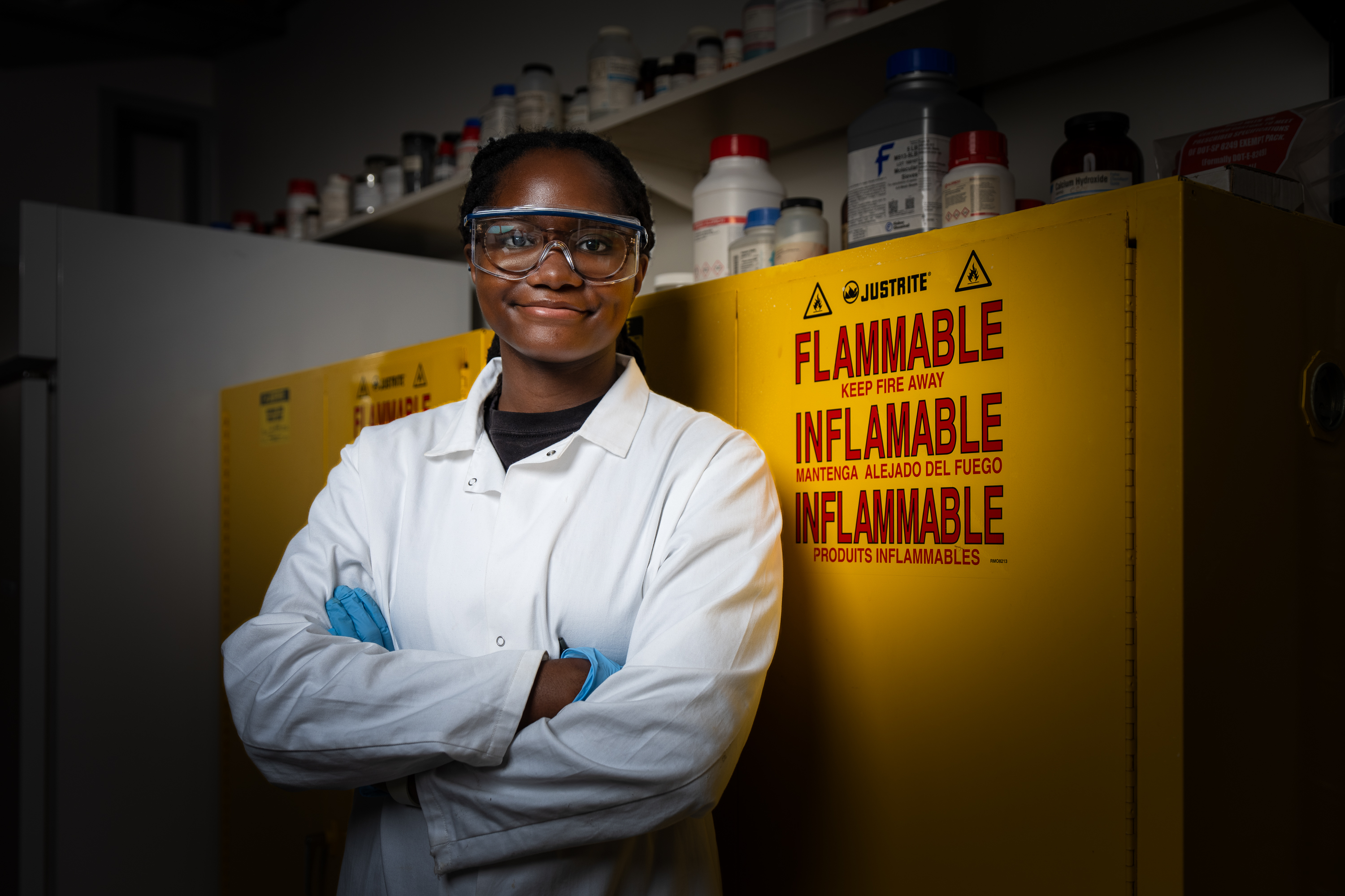 A researcher in a white lab coat, safety goggles, and gloves stands with arms crossed in front of yellow flammable-chemical storage cabinets lined with reagent bottles.