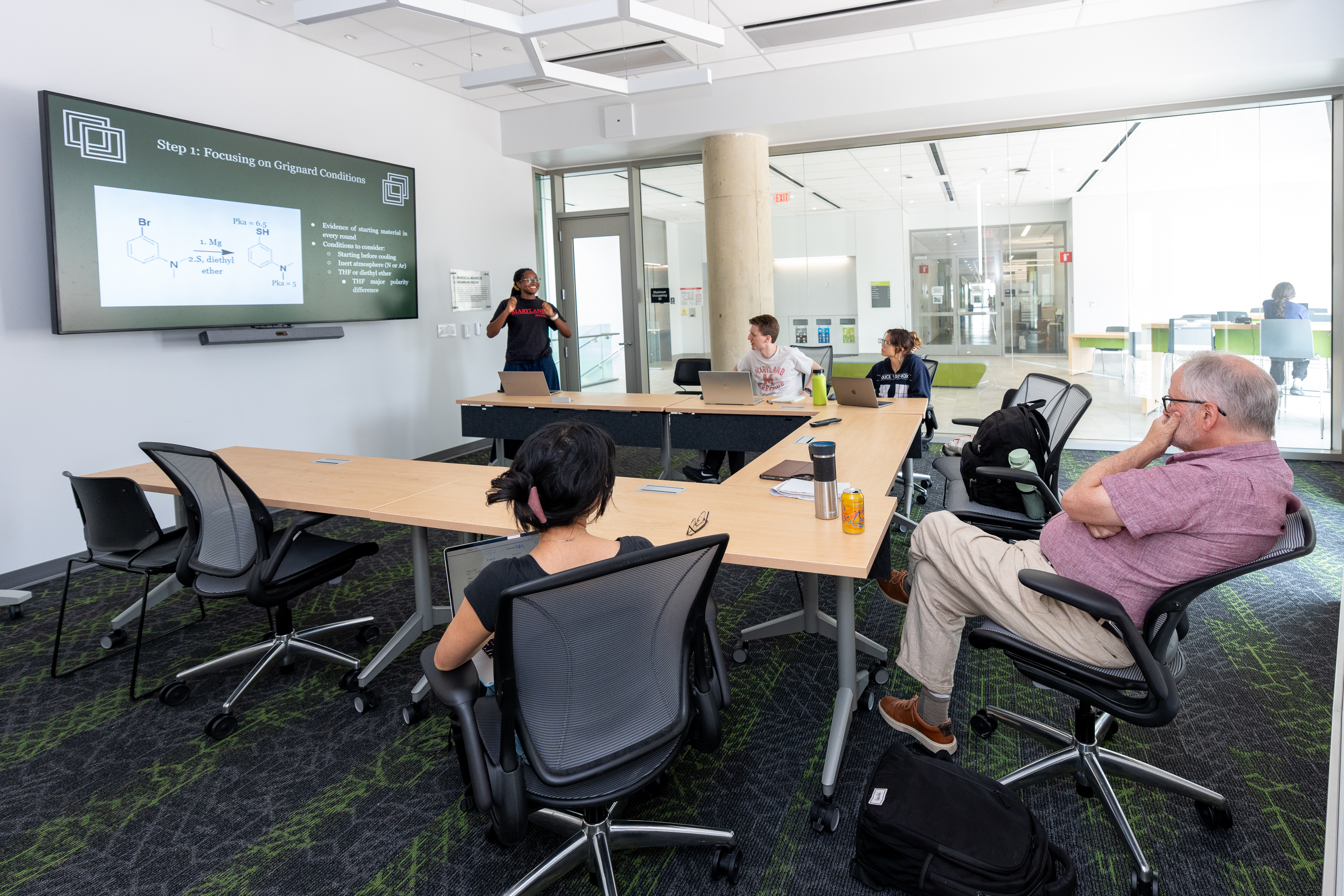 A researcher presents a slide on Grignard reaction conditions to colleagues seated around tables in a seminar room, with organic chemistry structures displayed on a large screen.