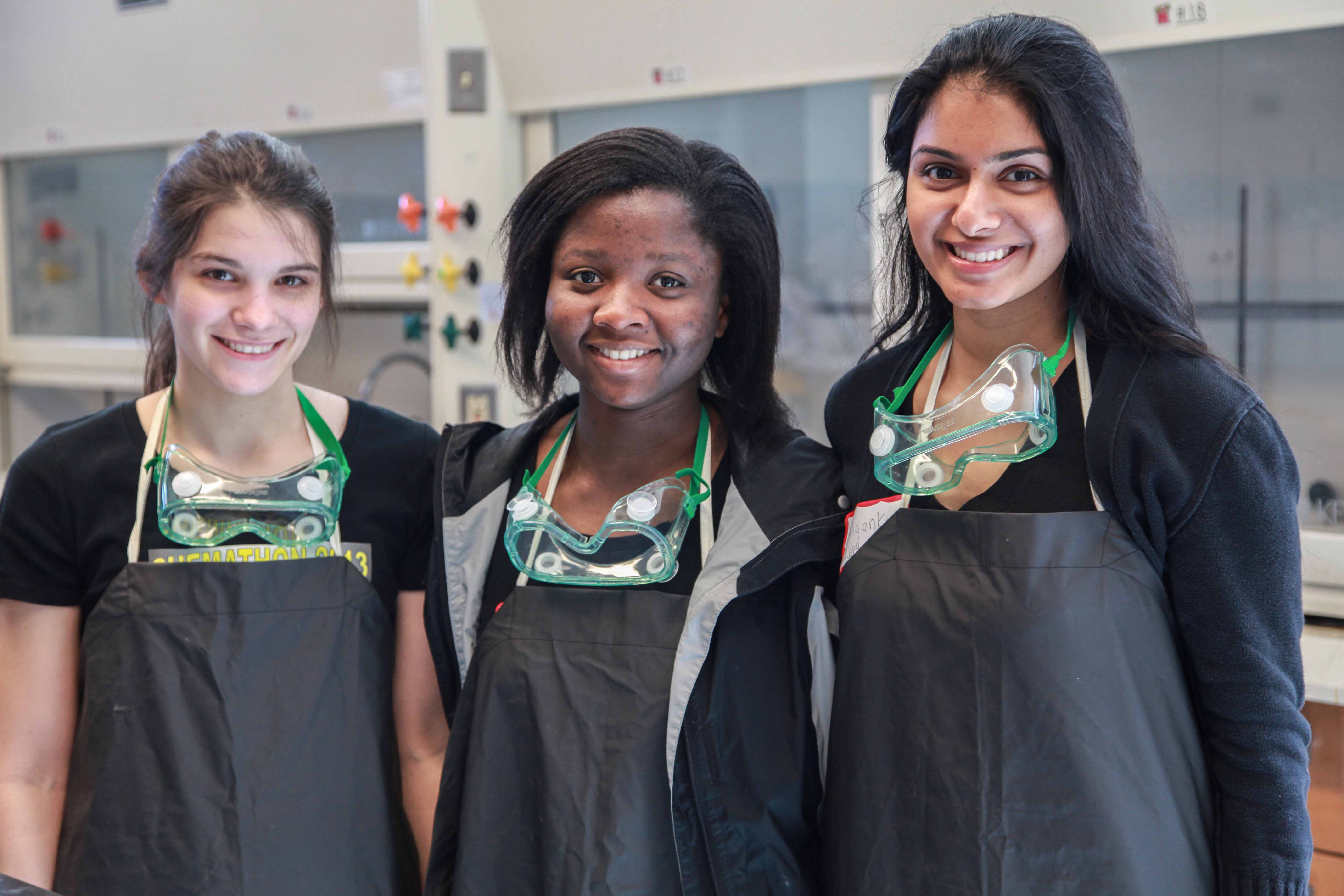 Three young women participating in Chemathon, wearing protective aprons and goggles.