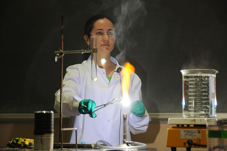 Lenea Stocker, dressed in a lab coat and goggles, using a burner for an experiment