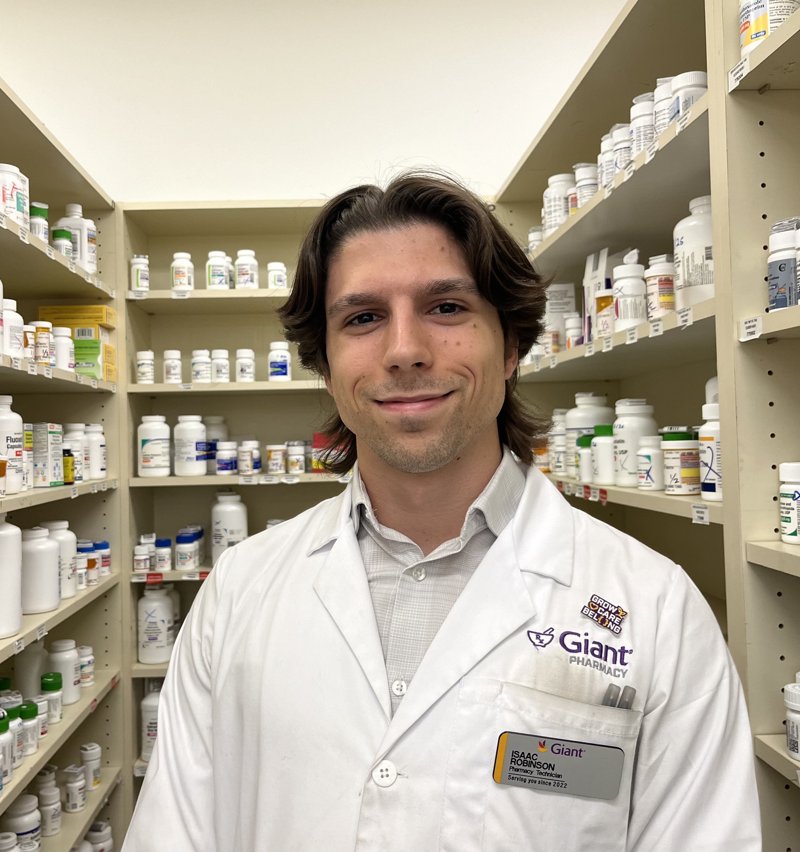 Isaac Robinson in a white lab coat stands in front of shelves of medication at Giant Pharmacy.