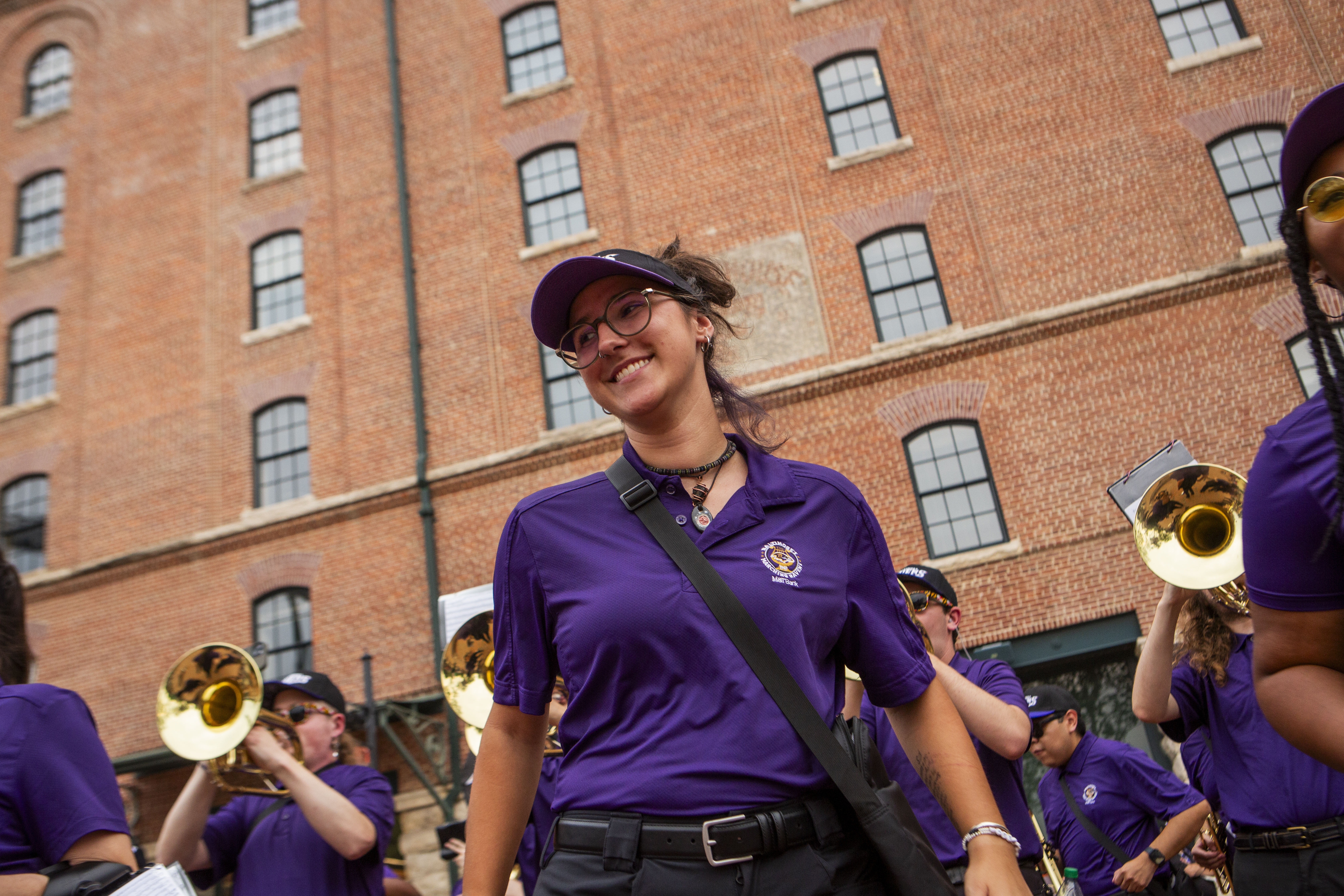 A smiling young woman walks outdoors with a marching band wearing a purple polo shirt, dark visor, and glasses, set against a large multi-story red brick building with tall windows. Other band members in matching purple shirts carrying brass instruments are visible around her.