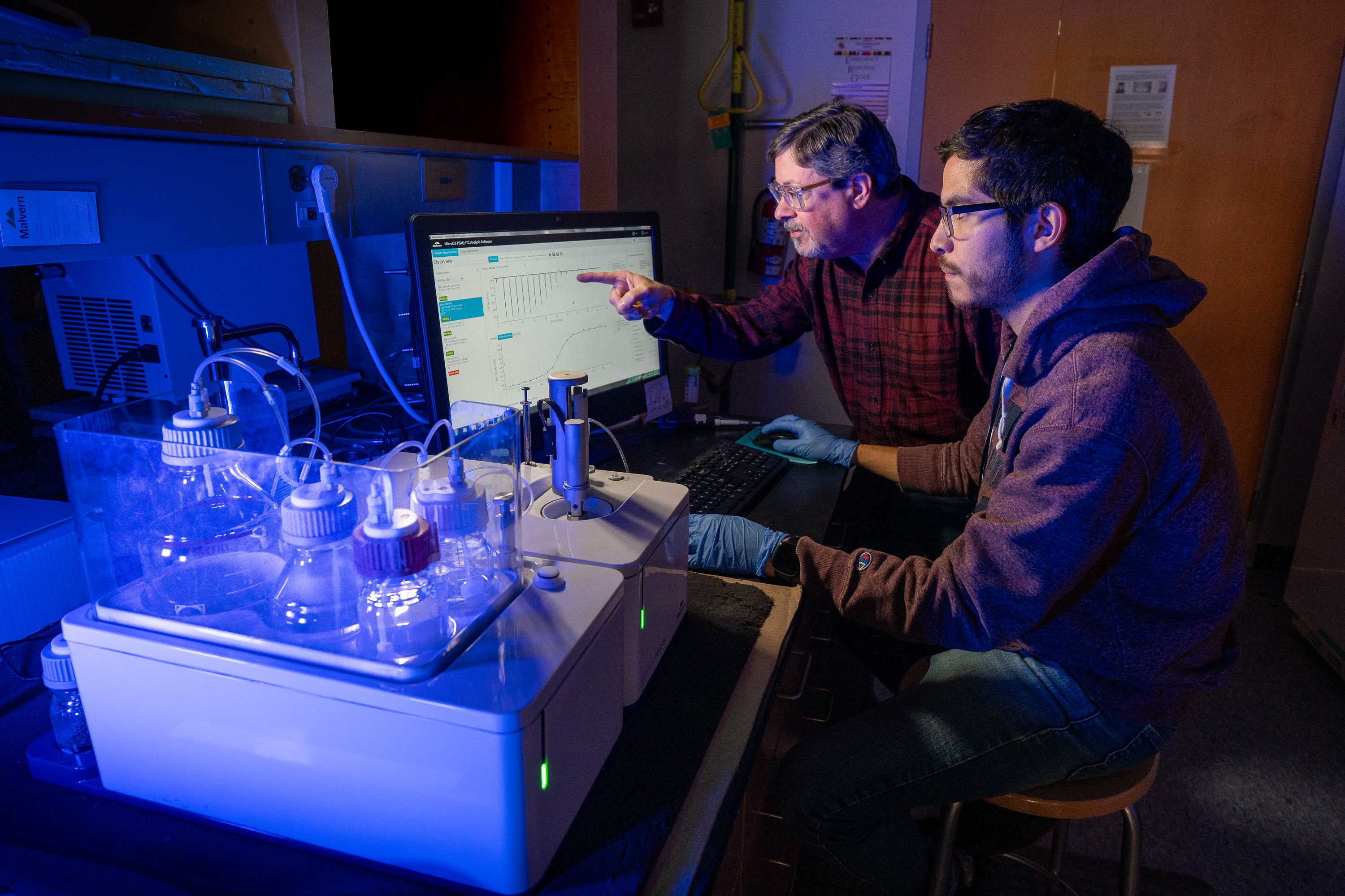 Two researchers work on a computer in a dimply lit room. One older man wears a checkered red button-down shirt, while a younger man wears a maroon hoodie. Both wear glasses. In front of them are beakers and flasks illuminated in blue light.