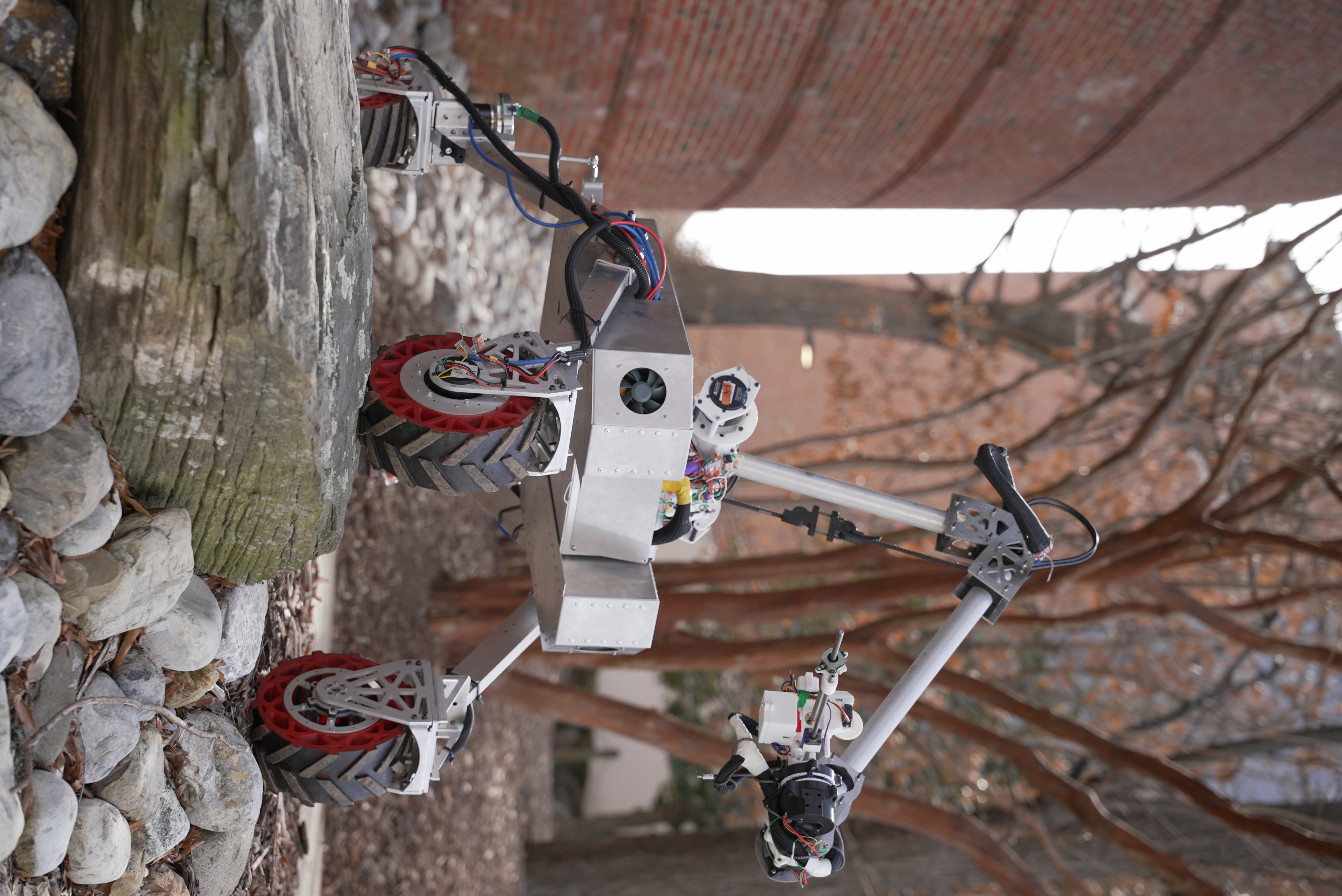 A small four-wheeled robotic rover with a mounted mechanical arm drives over rocks outdoors, near a brick building and leafless trees.