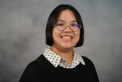 A young woman in a black-and-white polka dot button-down shirt and a black sweater smiles in front of a mottled gray backdrop.