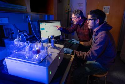 Two researchers work on a computer in a dimply lit room. One older man wears a checkered red button-down shirt, while a younger man wears a maroon hoodie. Both wear glasses. In front of them are beakers and flasks illuminated in blue light.