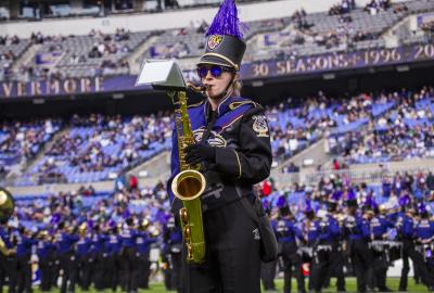A member of a marching band stands on a field holding a tenor saxophone, wearing a detailed purple and black uniform with gold trim, a black hat topped with a tall purple feather plume, and dark sunglasses. The blurred background shows a crowded stadium seating area under a "30 SEASONS" sign and other band members lined up on the field.