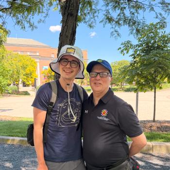 Two men stand outside, smiling on a sunny day. The one on the left wears gray shorts, a navy t-shirt, and a light gray bucket hat. The one on the right wears black jeans, a black University of Maryland promo, and a blue baseball cap.