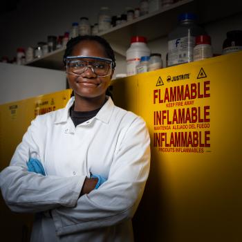 A researcher in a white lab coat, safety goggles, and gloves stands with arms crossed in front of yellow flammable-chemical storage cabinets lined with reagent bottles.