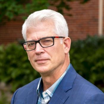 A man in black glasses and a blue suit over a blue plaid shirt stands in front of blurred greenery and bricks.