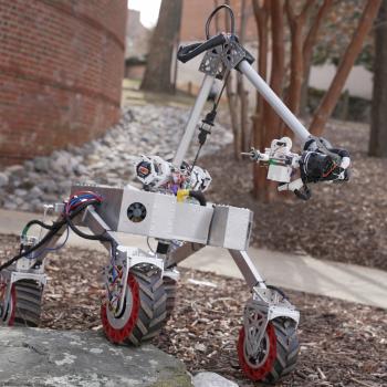 A small four-wheeled robotic rover with a mounted mechanical arm drives over rocks outdoors, near a brick building and leafless trees.