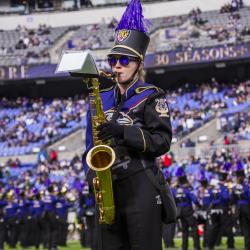 A member of a marching band stands on a field holding a tenor saxophone, wearing a detailed purple and black uniform with gold trim, a black hat topped with a tall purple feather plume, and dark sunglasses. The blurred background shows a crowded stadium seating area under a "30 SEASONS" sign and other band members lined up on the field.